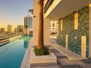 A modern pool area with a clear view of the city skyline, a palm tree, and a tiled wall. - Modern Chic Studio Apartment - JVC Dubai (Dubai)