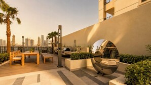 An outdoor patio with a wooden table and chairs, a grill, and a decorative mirror. - Modern Chic Studio Apartment - JVC Dubai (Dubai)