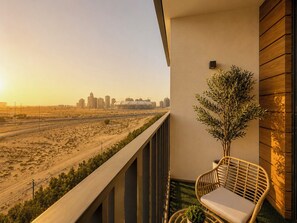 A balcony with a view of a desert landscape and city skyline, a wicker chair, and a small potted plant. - Modern Chic Studio Apartment - JVC Dubai (Dubai)