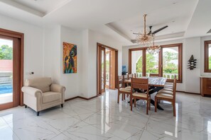 A spacious dining area with a wooden table and chairs, a ceiling fan, and a large window with a view of greenery. - Villa Branca Diani: Spacious 5-bedroom luxury with personalized service. (Diani Beach)