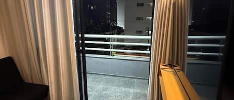 A hotel room with a sliding glass door leading to an outdoor balcony, a dark-colored sofa, a wooden side table with a remote control, and a large mirror.