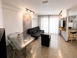 A modern living room with a glass dining table, a black sofa, and a kitchen area with a TV. - 2-bedroom apartment in perfect Fortaleza with refreshing AC (Fortaleza)