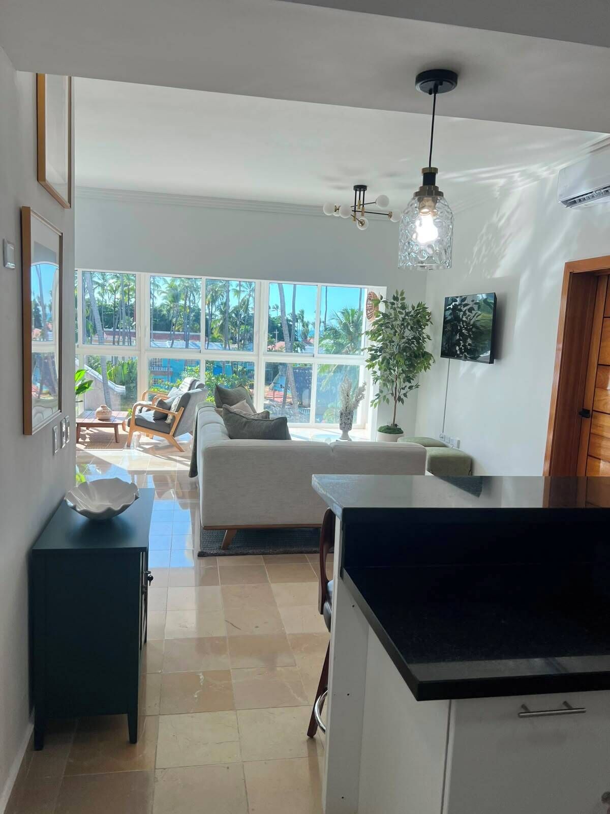 A modern kitchen with a large island, a dining area with a sofa and chairs, and a view of palm trees through large windows.