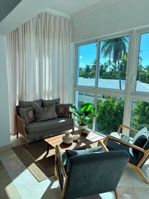 A modern living room with a sofa, coffee table, and chairs, large windows with a view of palm trees. - Beachfront Penthouse w/ Rooftop, Pools & Views (Punta Cana)