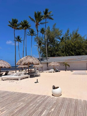 A beachside area with thatched umbrellas, lounge chairs, and a wooden deck. - Beachfront Penthouse w/ Rooftop, Pools & Views (Punta Cana)