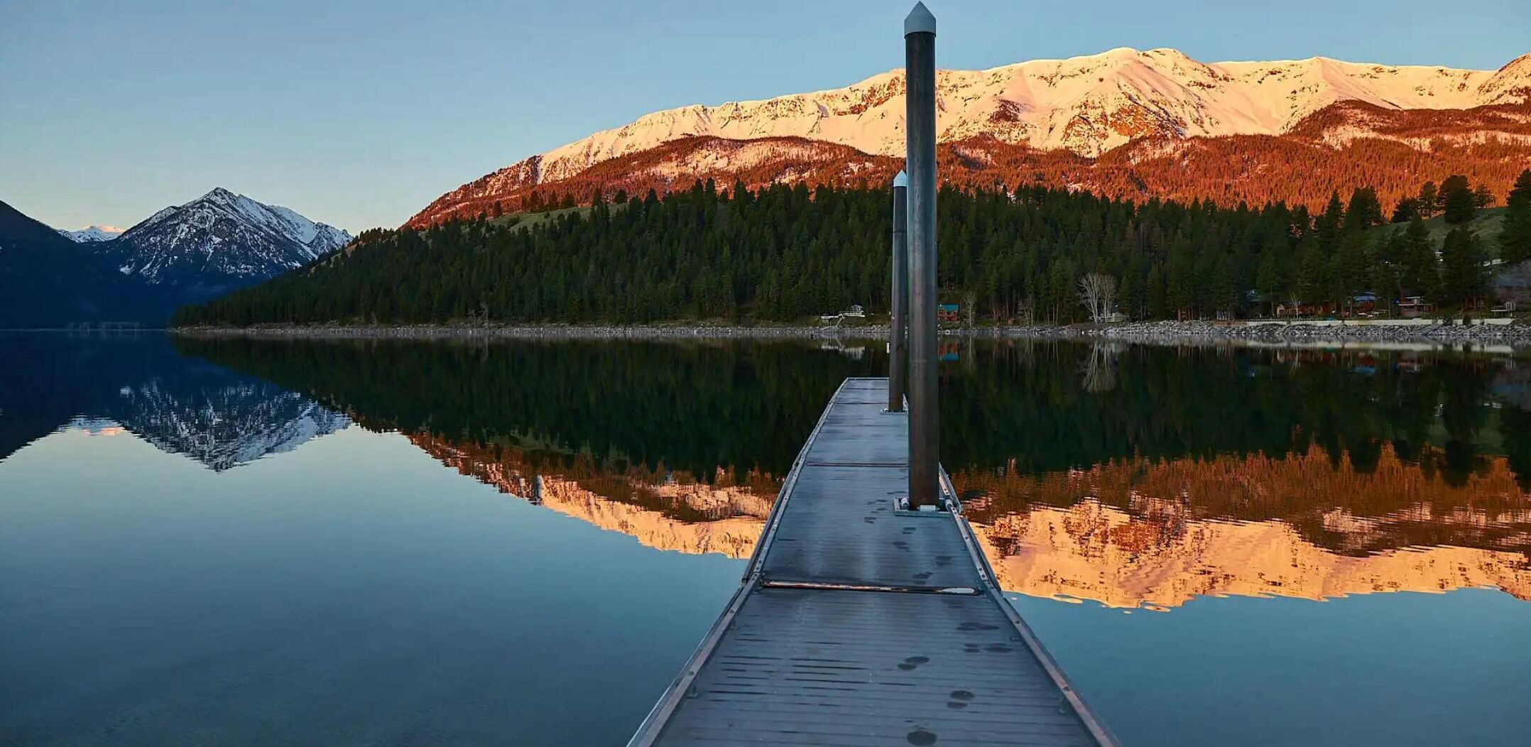 A wooden dock extending into a calm lake with a mountain range reflected in the water.