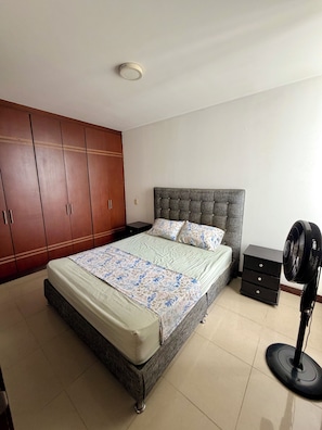 Ein Schlafzimmer mit Bett, Kleiderschrank, Nachttisch und Standventilator.