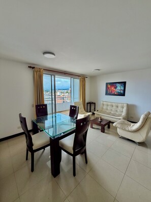 A modern living room with a glass dining table, two sofas, and a large window with curtains. - Apto Lili - Cali - Wifi - Familiar, Piscina (Cali)