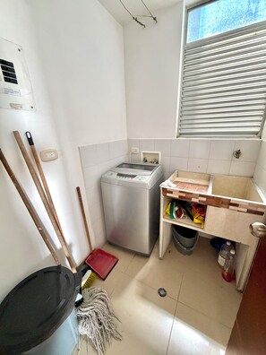 A small laundry room with a washing machine, a mop, a broom, and a trash bin. - Apto Lili - Cali - Wifi - Familiar, Piscina (Cali)