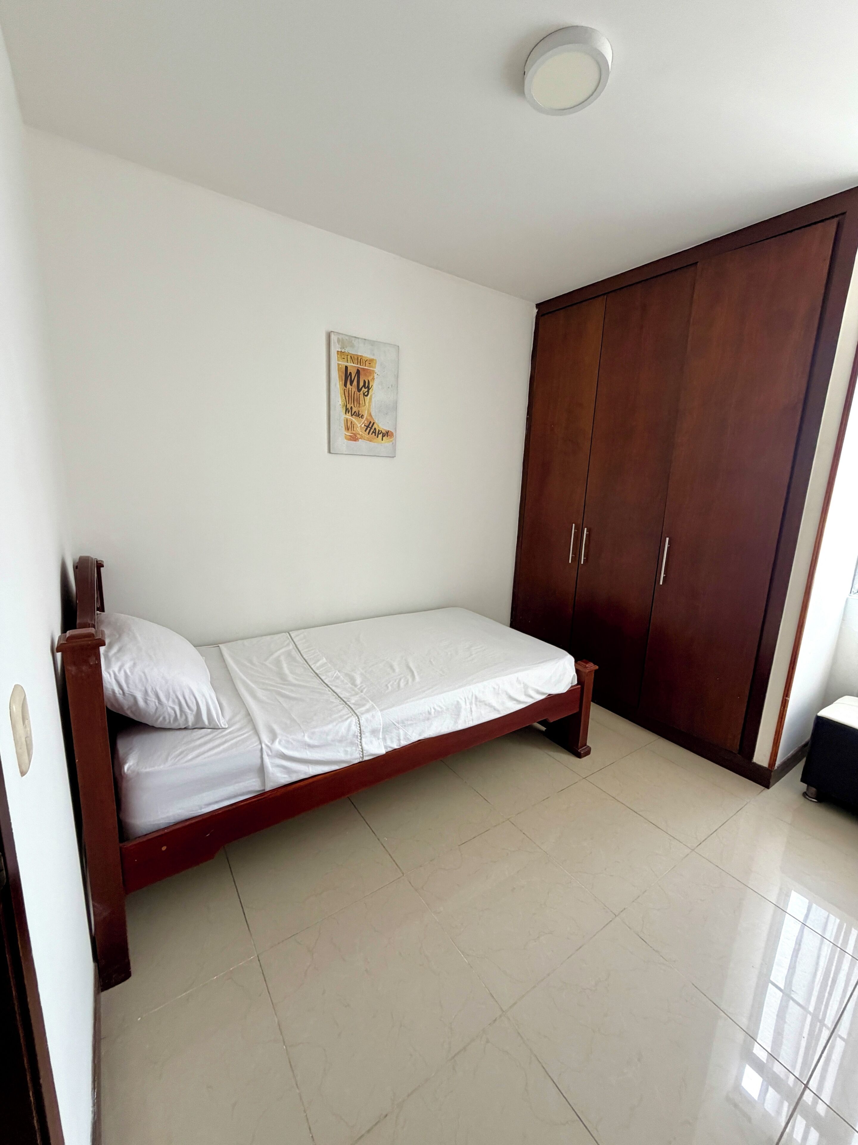 A single bed with a wooden frame and white bedding in a room with a tiled floor and a wardrobe.