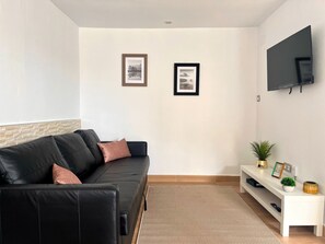 A modern living room with a black leather sofa, two framed pictures on the wall, a flat-screen TV, and a white side table with decorative items.