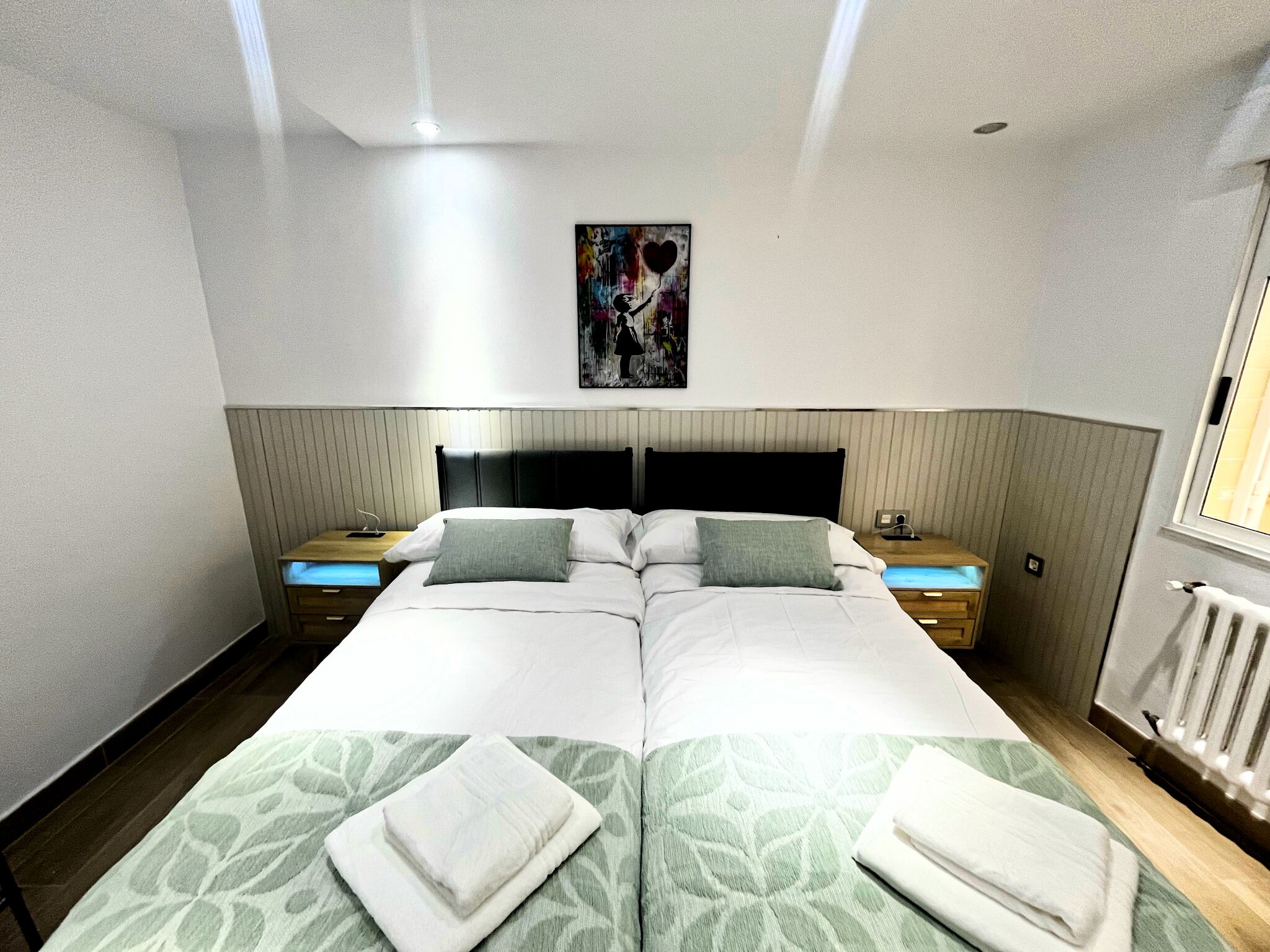 A double bed with a green patterned bedspread, two nightstands with lamps, a wall-mounted painting, and a window with blinds.