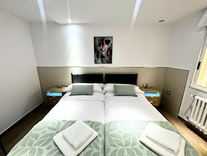 A double bed with a green patterned bedspread, two nightstands with lamps, a wall-mounted painting, and a window with blinds.
