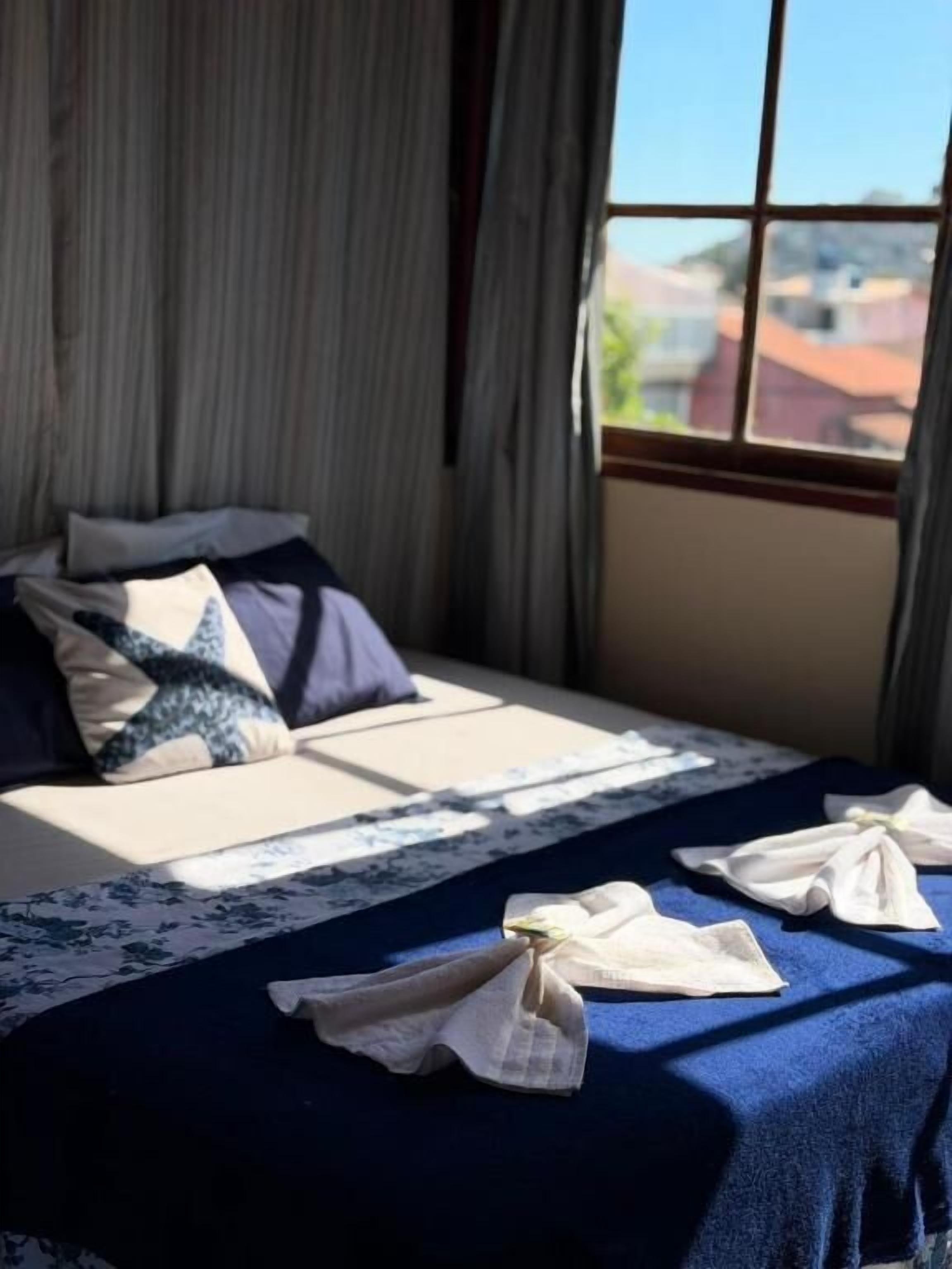 Un lit avec une couvre-lit bleue et des oreillers, une fenêtre avec des rideaux et une vue sur les bâtiments à l’extérieur.