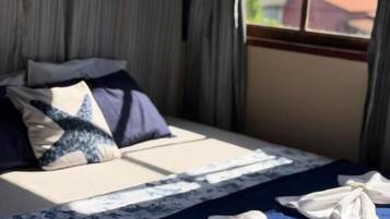 Un lit avec une couvre-lit bleue et des oreillers, une fenêtre avec des rideaux et une vue sur les bâtiments à l’extérieur.