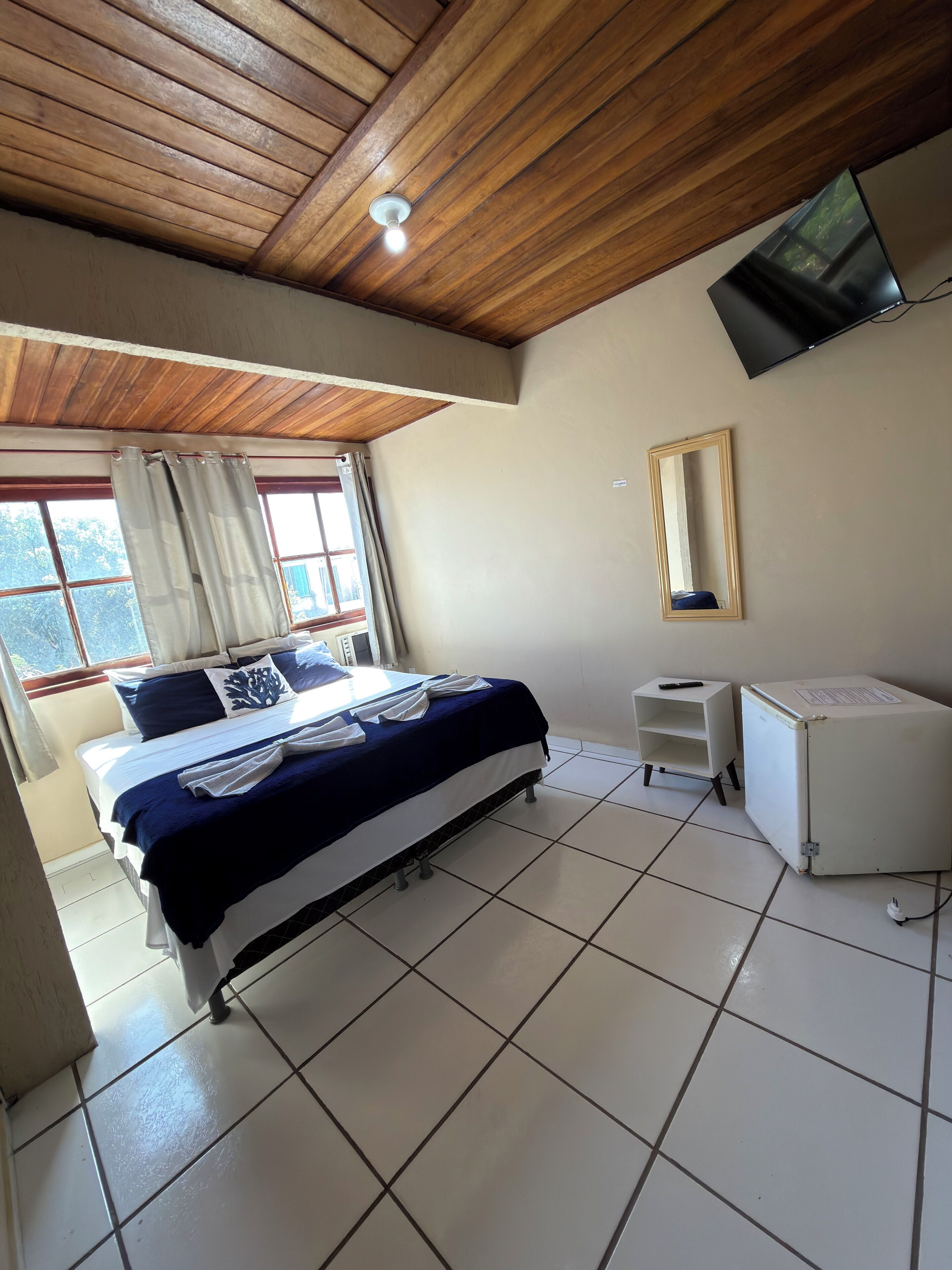 A bedroom with a bed, a television mounted on the wall, a small white cabinet, and a window with curtains.