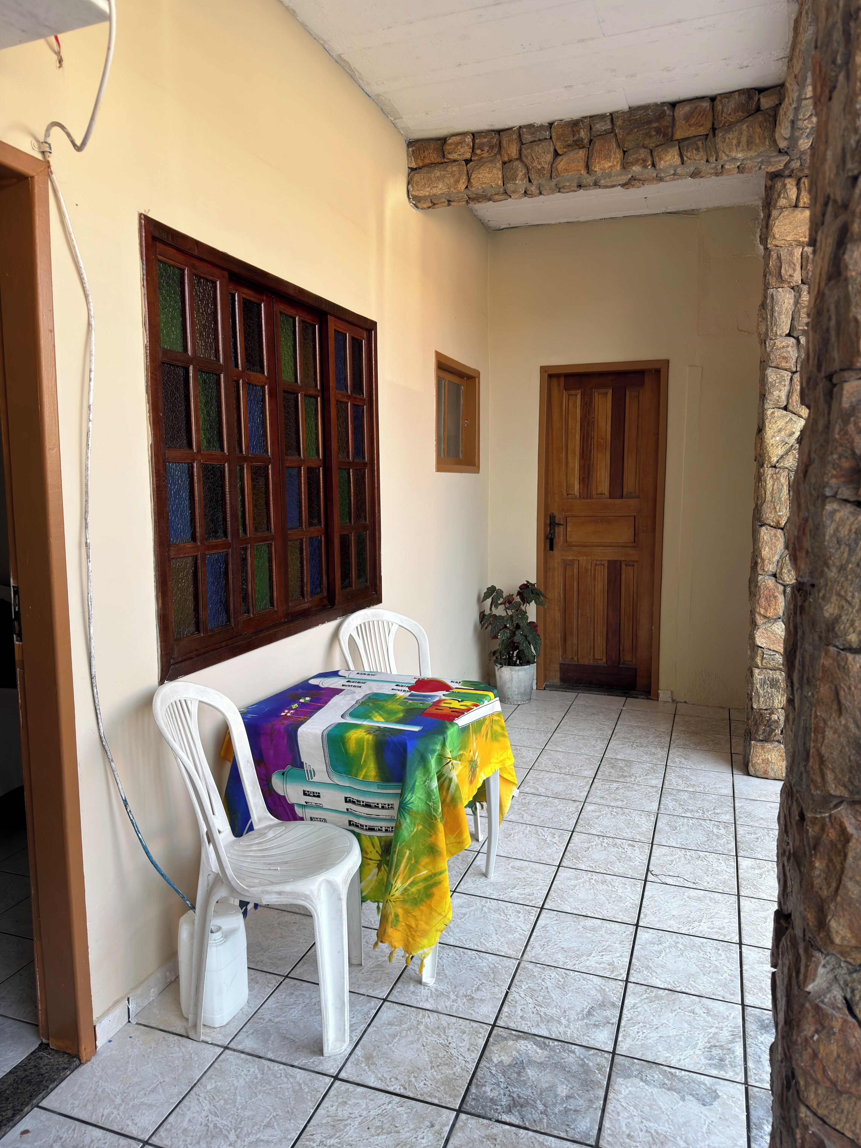 A room with a tiled floor, a table with a colorful tablecloth, two white plastic chairs, a wooden door, and a stone wall.
