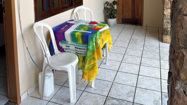 Una habitación con piso de baldosas, una mesa con mantel colorido, dos sillas de plástico blanco, una puerta de madera y una pared de piedra.