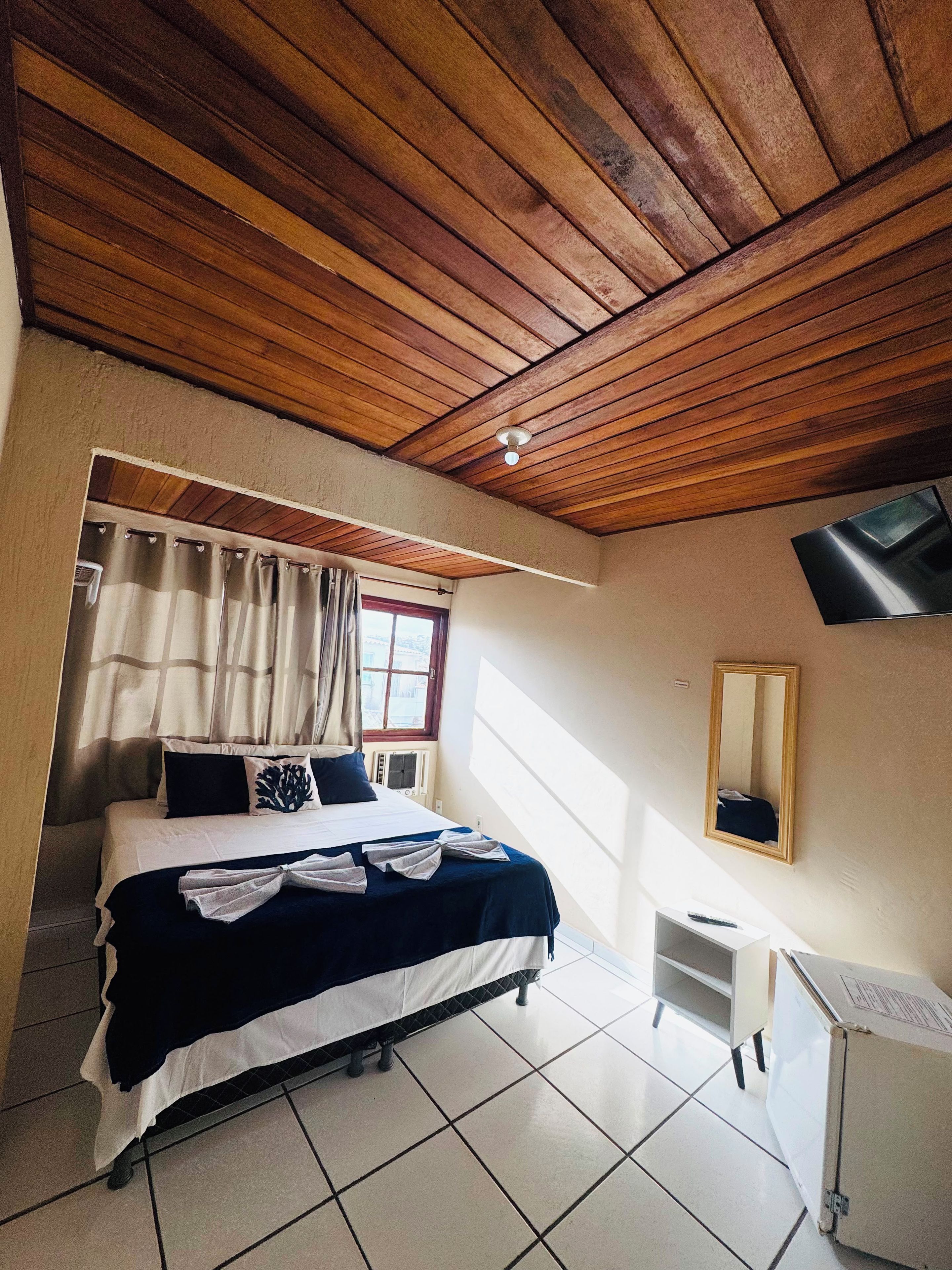 A hotel room with a wooden ceiling, a bed with a dark blue bedspread, a white bedside table, a small white cabinet, a window with curtains, and a wall-mounted TV.