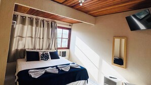 Habitación de hotel con techo de madera, cama con colcha azul oscuro, mesita de noche blanca, armario blanco pequeño, ventana con cortinas y televisor montado en la pared.