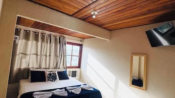 A hotel room with a wooden ceiling, a bed with a dark blue bedspread, a white bedside table, a small white cabinet, a window with curtains, and a wall-mounted TV.