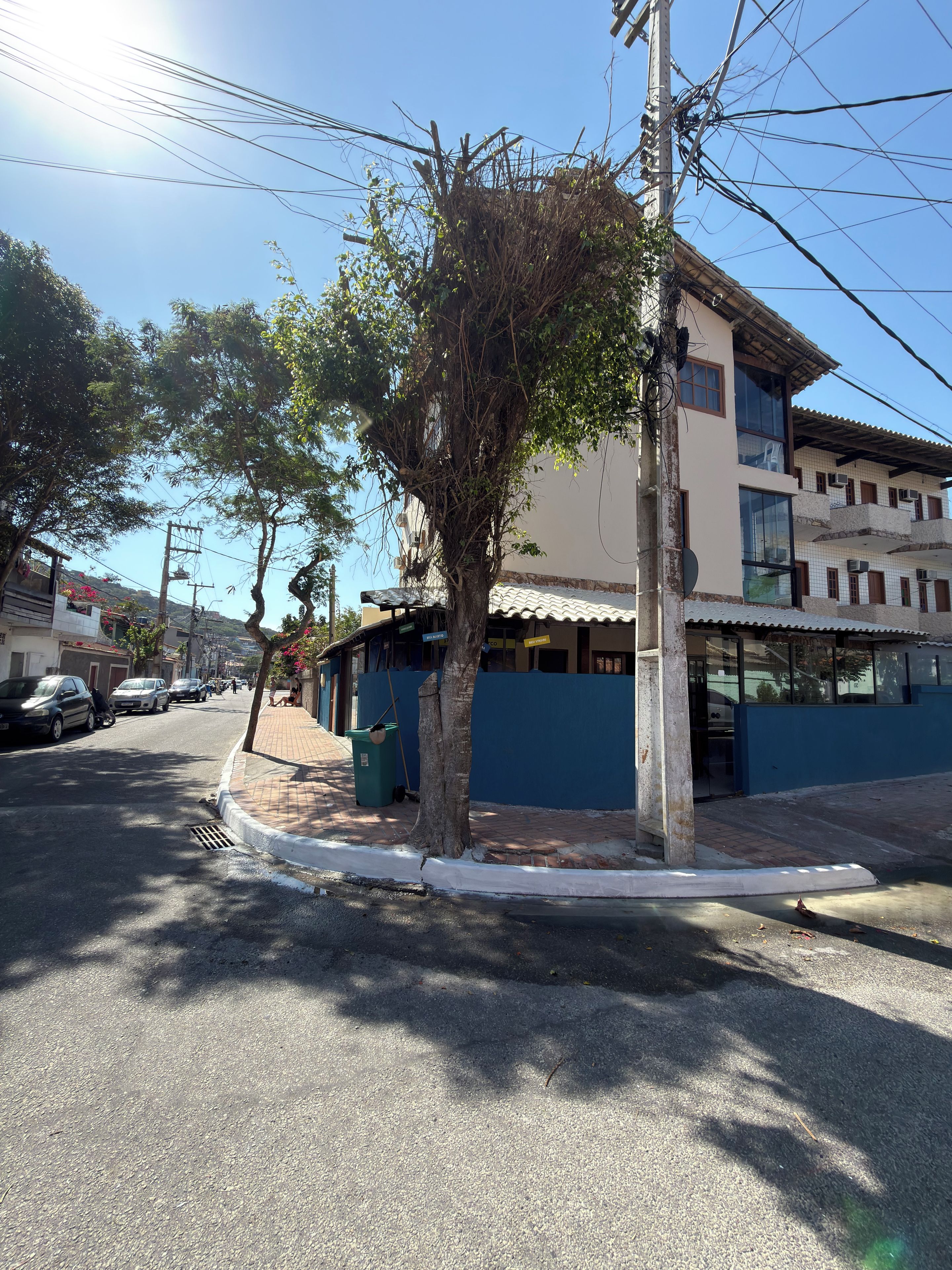 Vista da rua com um edifício com varanda, uma árvore e carros estacionados.