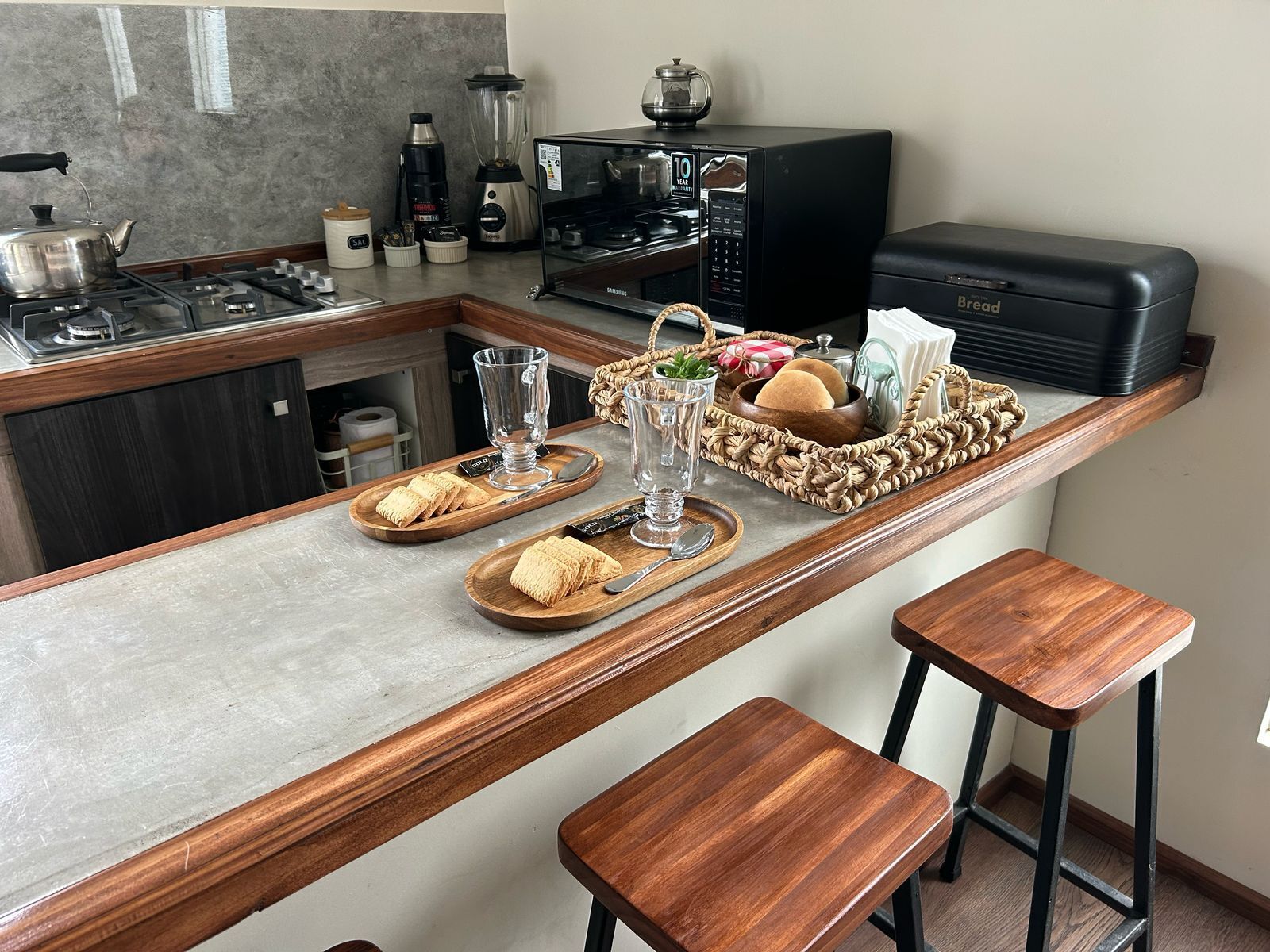 A kitchen with a built-in microwave, a kettle, and a coffee maker. There are two wooden stools and a tray with bread and butter.