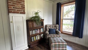 A room with a brick wall, a bookshelf, a plaid chair, a window with curtains, and a wooden floor.
