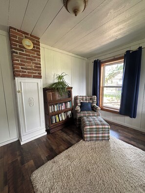 A room with a brick wall, a bookshelf, a plaid chair, a window with curtains, and a wooden floor.
