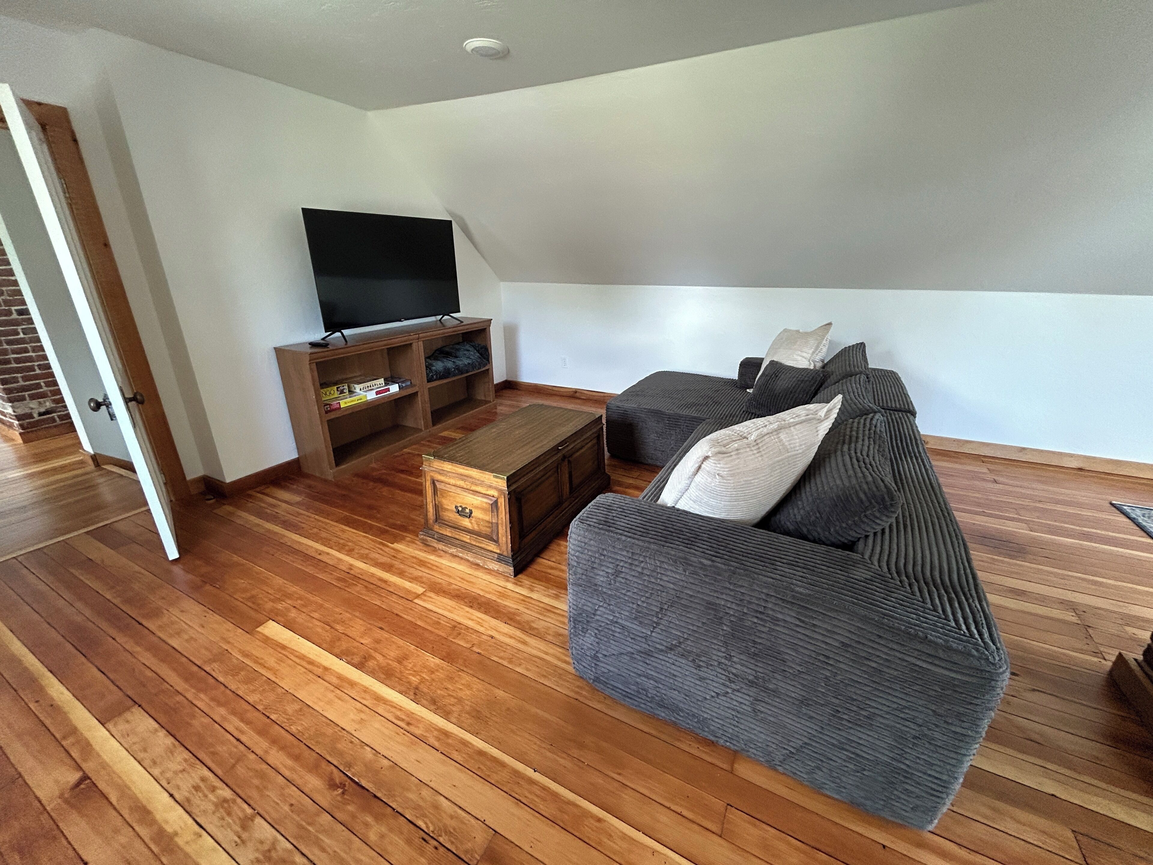 Een woonkamer met een houten vloer, een grijze bank, een televisie op een houten dressoir, en een klein houten bijzettafeltje.