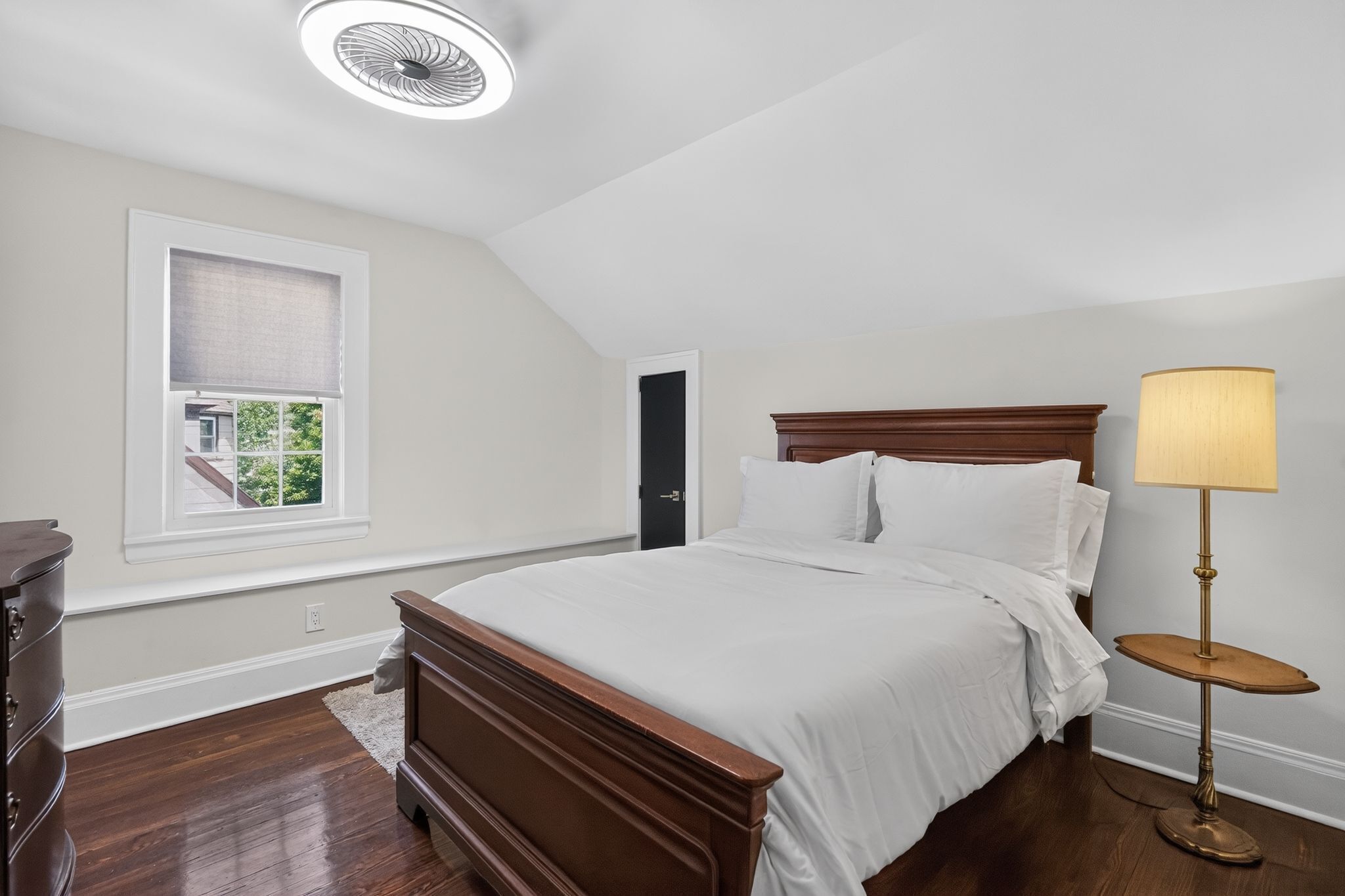 Une chambre à coucher comprenant un lit, une table de chevet, une lampe, une fenêtre avec des stores et une porte.