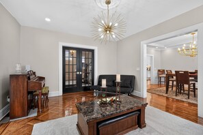 A spacious living room with a wooden piano, a black sofa, a marble coffee table, and a dining area with wooden chairs and a chandelier. - Historic Forest Hill Single Family Near NYC (Newark)