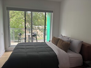 A bedroom with a large window, a bed with a dark quilt, and a view of a balcony with chairs. - Le Diamant Bleu in Quebec City ~GYM~GARAGE~Downtown (Québec)