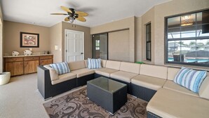 A living room with a sectional sofa, a glass coffee table, a ceiling fan, a kitchen area with wooden cabinets, and a window with a view of a swimming pool. - Peaceful, Canal Front Home w/ Heated Pool - Surfside Beach House (Apollo Beach)