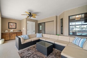 A living room with a sectional sofa, a glass coffee table, a ceiling fan, a kitchen area with wooden cabinets, and a window with a view of a swimming pool. - Peaceful, Canal Front Home w/ Heated Pool - Surfside Beach House (Apollo Beach)