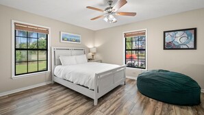 A bedroom with a bed, a ceiling fan, two windows, a bedside table, and a green bean bag chair. - Peaceful, Canal Front Home w/ Heated Pool - Surfside Beach House (Apollo Beach)