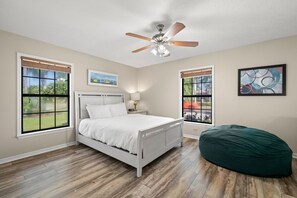 A bedroom with a bed, a ceiling fan, two windows, a bedside table, and a green bean bag chair. - Peaceful, Canal Front Home w/ Heated Pool - Surfside Beach House (Apollo Beach)