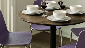 Mesa de jantar com tigelas brancas e copos de vinho, rodeada por cadeiras roxas.