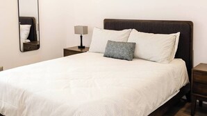 Uma cama bem arrumada com colcha listrada, uma mesa de cabeceira com abajur e um espelho fixado na parede.
