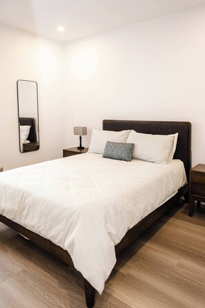A neatly made bed with a striped bedspread, a bedside table with a lamp, and a wall-mounted mirror. - New Vineyard Cottage in Niagara Wine Country | 2BR | Pet Friendly (Lincoln)