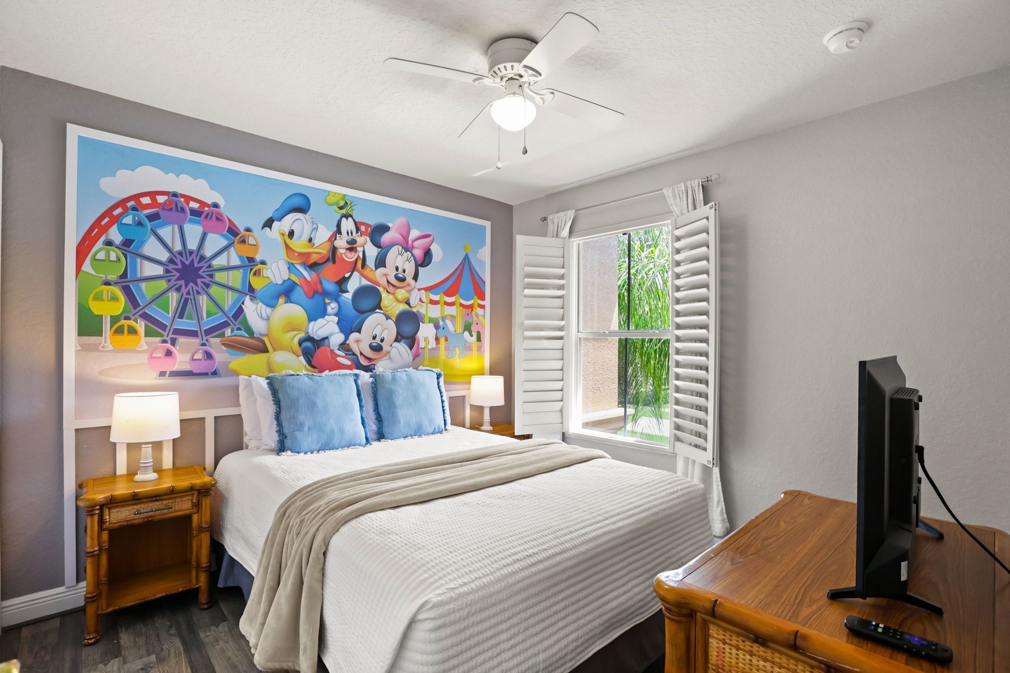Habitación de hotel con un gran mural de personajes animados, una cama con ropa blanca, una mesita de noche de madera, una lámpara, una ventana con contraventanas y un televisor sobre un mueble de madera.