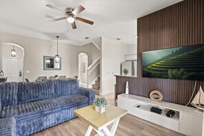 A modern living room with a blue sofa, a wooden coffee table, a TV mounted on a wooden wall, and a ceiling fan. - 4 BD/ 3 BA/ Regal Palms (3214 CA) (Davenport)