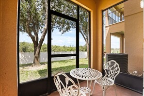 A covered outdoor seating area with a table and two chairs, a view of trees and a white fence, and a building with a balcony in the background. - 4 BD/ 3 BA/ Regal Palms (3214 CA) (Davenport)
