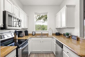 Cocina moderna con armarios blancos, encimera de granito, electrodomésticos de acero inoxidable y ventana con vistas a árboles.