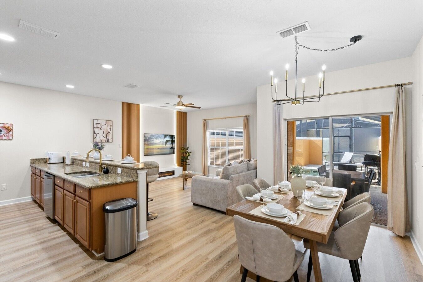 A modern kitchen with a central island, dining area with a table set for four, and a living room with a sofa and a view of the outdoors.