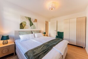 A modern bedroom with a bed, nightstands, a green chair, a wardrobe, and a wall art piece. - Ostseeglück Sassnitz - Stylish Apartment by the Sea (Sassnitz)