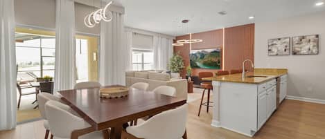 Una cocina moderna con una isla central, comedor con mesa y sillas de madera, y sala de estar con sofá y un ventanal grande.
