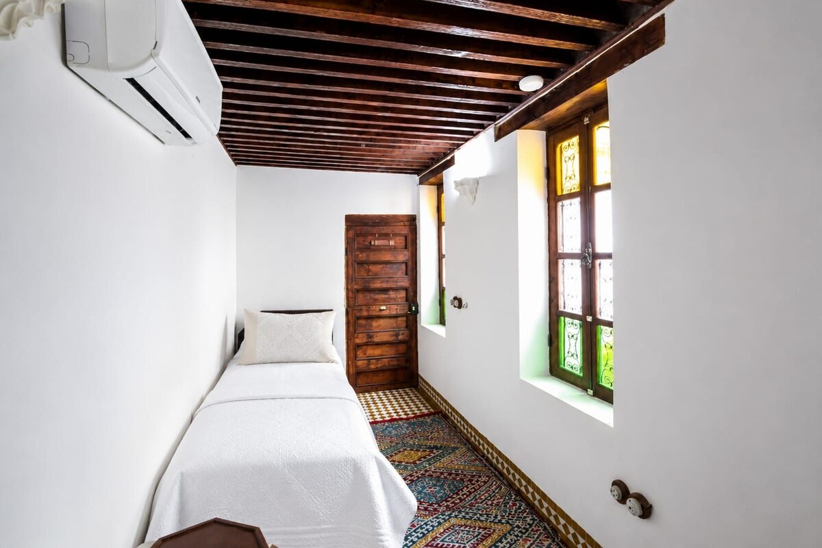 Un pasillo estrecho con una cama, una puerta de madera y un ventanal con vidrio verde.