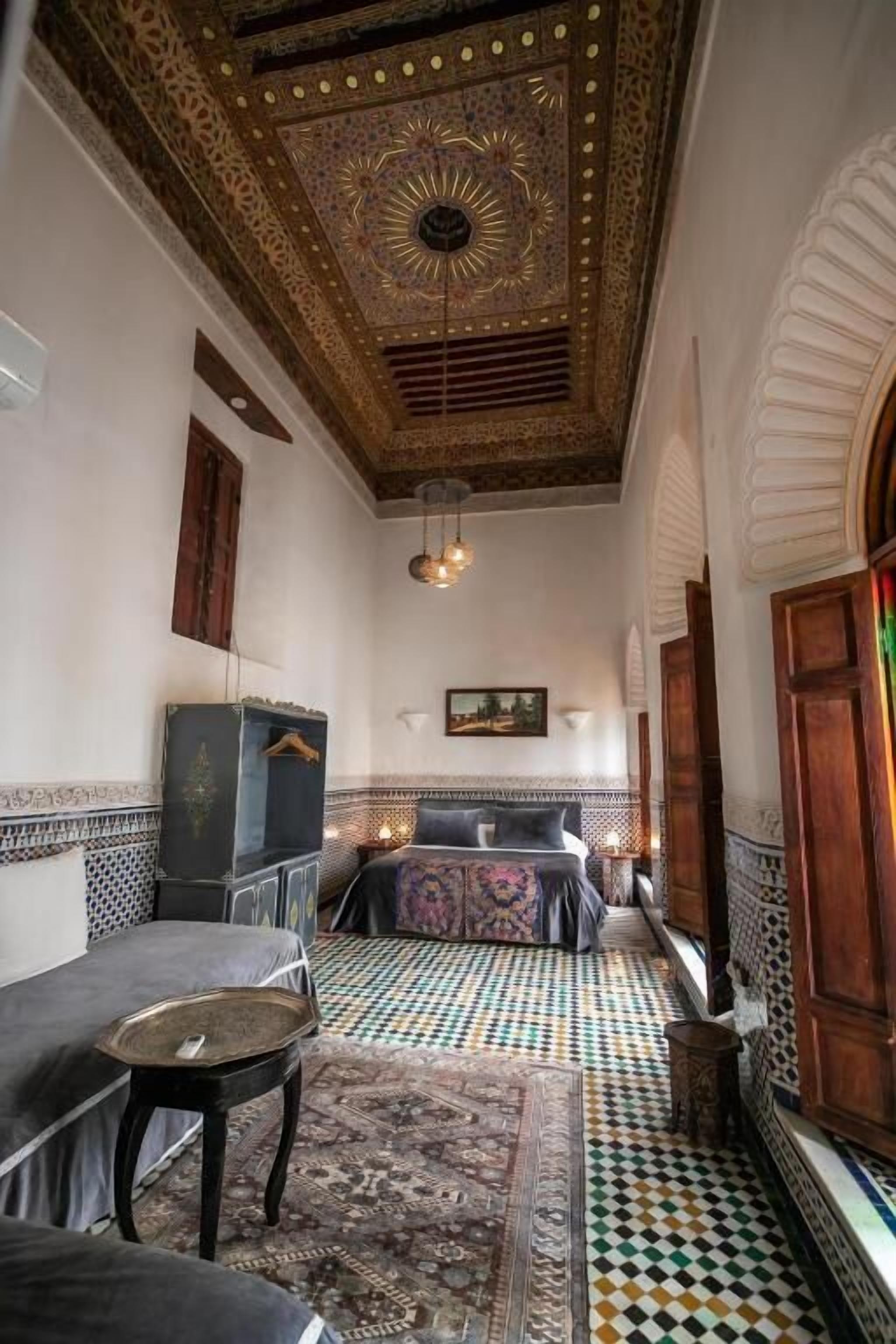 A room with a patterned floor, a bed with a patterned bedspread, a small table, and a decorative ceiling.