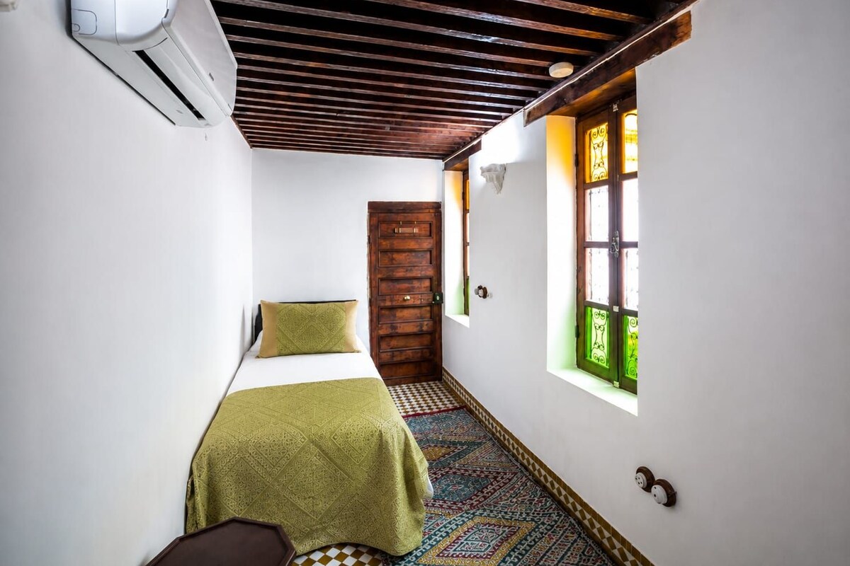 Una habitación estrecha con una cama, una puerta de madera y un ventanal con vidrios verdes.